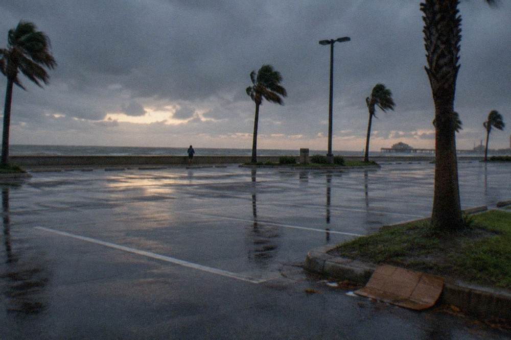 A wide retail parking lot at dusk with palm trees and a darkening sky over the seawall